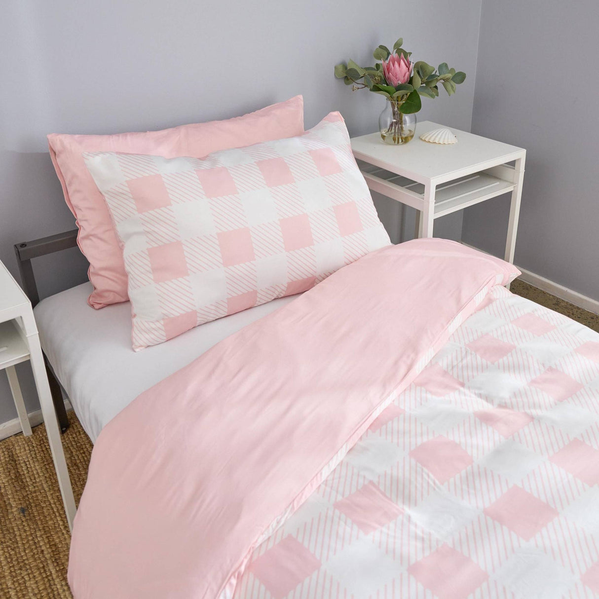 Pink and white checkered bedding set on a bed with a side table and plant in the background.