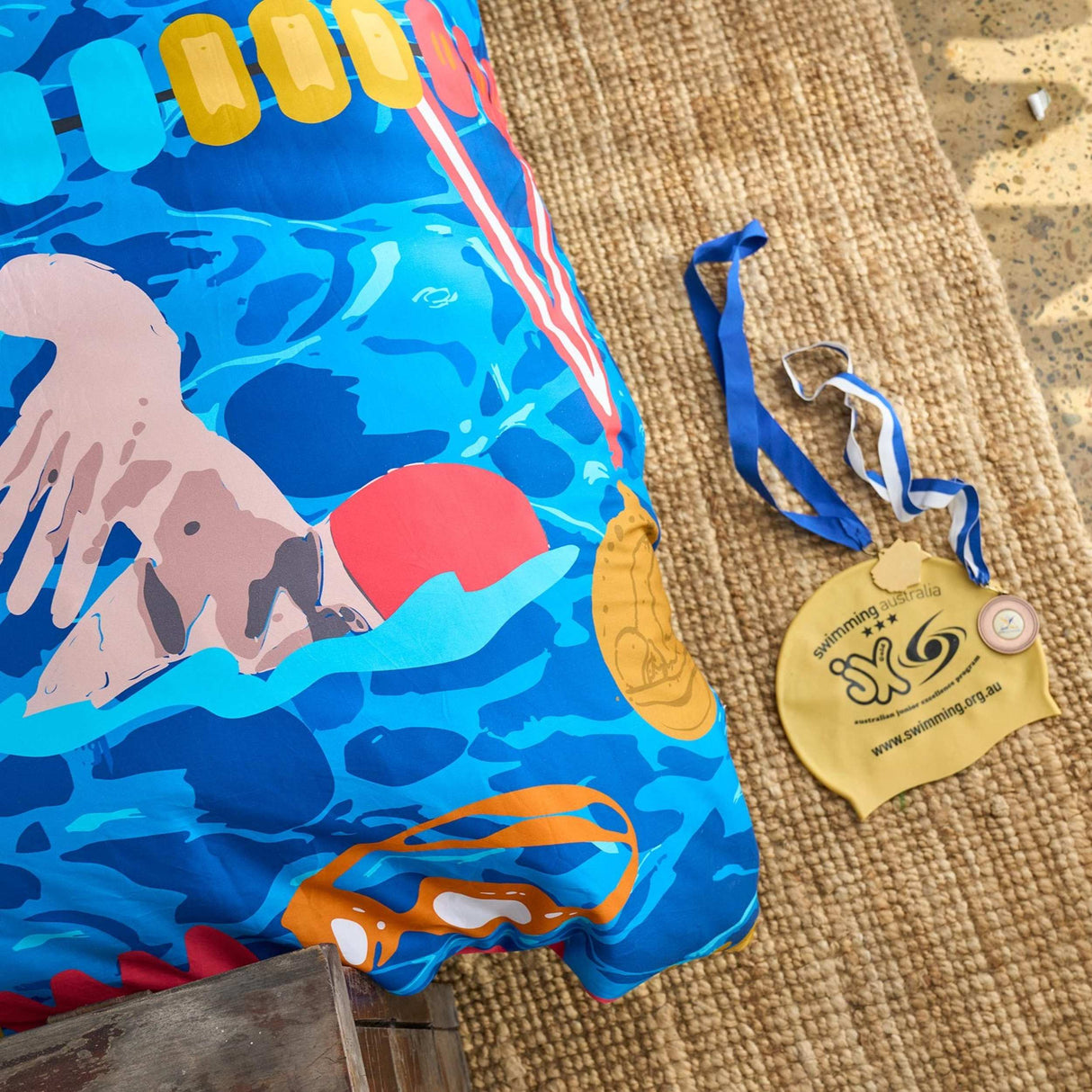 Detail of swimming themed quilt cover on bed with swimming cap and medal on floor next to it.
