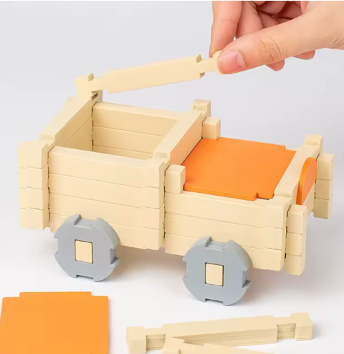 Hand building a wooden toy cart with orange seats and gray wheels on a white background