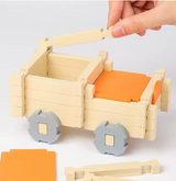 Hand building a wooden toy cart with orange seats and gray wheels on a white background